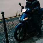 Motociclista de capacete conduz uma scooter por uma rua de paralelepípedos.