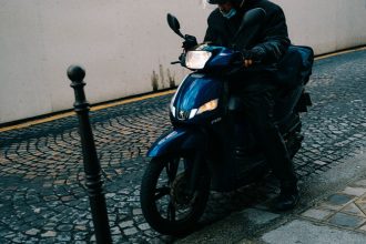 Motociclista de capacete conduz uma scooter por uma rua de paralelepípedos.
