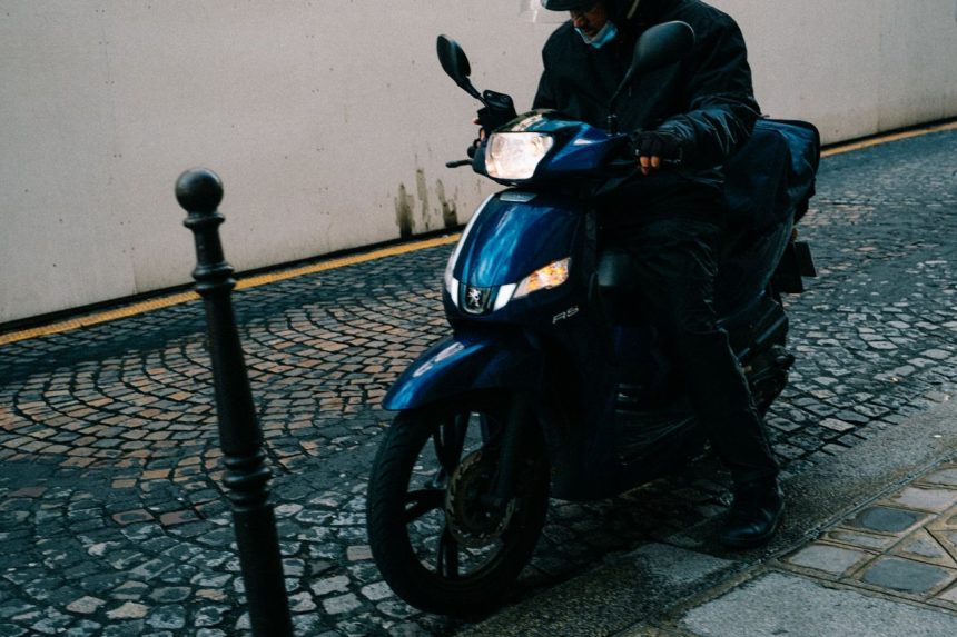 Motociclista de capacete conduz uma scooter por uma rua de paralelepípedos.