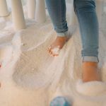 Criança brinca descalça em uma área interna de recreação com areia e brinquedos coloridos.